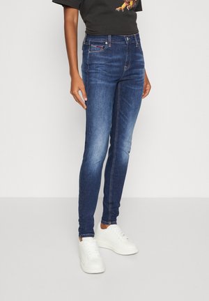 Femme portant un jean bleu foncé ajusté, des baskets blanches et un t-shirt noir, debout devant un fond clair uni.