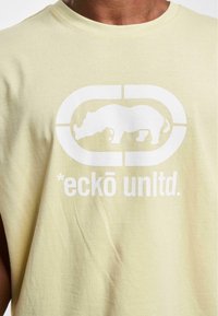 Lys gul ærmeløs t-shirt lavet af bomuld med et hvidt grafisk motiv af en næsehorn og teksten "*eckō unlt.d." trykt nedenunder.