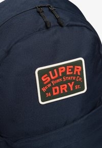 Mochila de tela azul marino con un parche que tiene texto rojo y verde que dice "SUPER DRY," un diseño texturizado y doble cremallera.