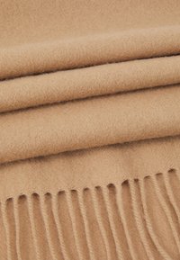 Couverture en laine beige à texture douce, avec des bords frangés et des plis superposés, présentant une couleur chaude et uniforme.