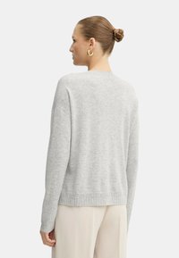 Donna con un bun intrecciato che indossa orecchini a cerchio dorati, un maglione di maglia grigio chiaro e pantaloni color crema, vista di spalle su uno sfondo neutro.
