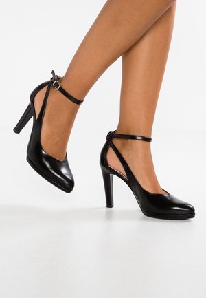 Chaussures à talons hauts en cuir verni noir avec bride à la cheville et côtés ouverts. Design à bout rond avec une texture brillante et un talon fin et effilé.