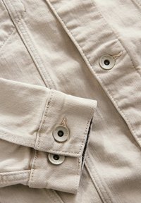 Lichtbeige denimjas met een opgerolde manchette en metalen knopen met gegraveerde merknaam. Prominente stikseldetails zichtbaar.
