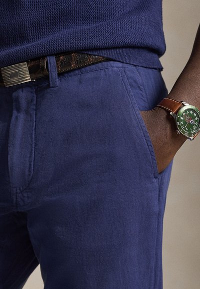 Marineblauwe broek van een gestructureerde stof, met voorzakken en een bruine leren riem. Aan de pols wordt een groen horloge gedragen.