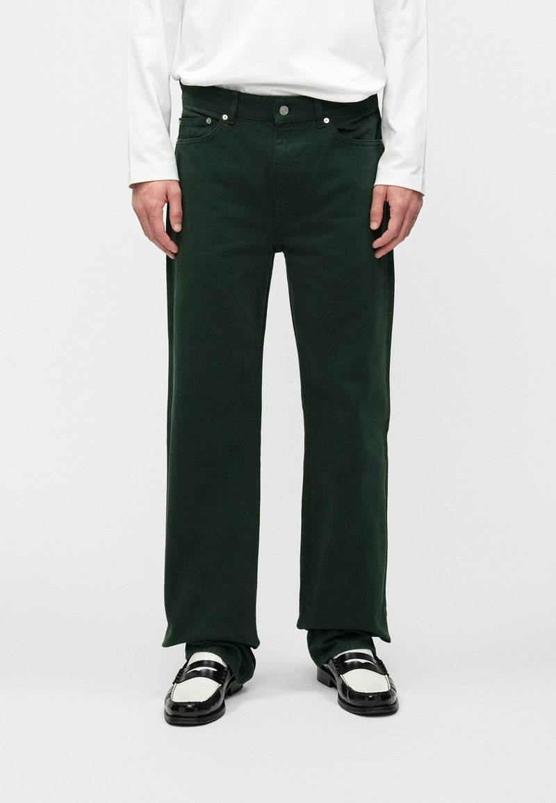 Pantalons en coton verts avec une coupe droite, dotés de poches avant, de boutons visibles et d'une texture lisse. Assortis avec des mocassins noir et blanc.