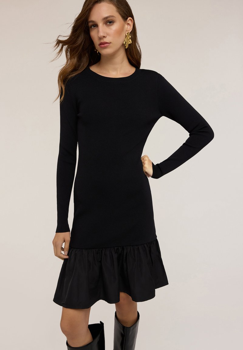 Abito in maglia nero con maniche lunghe, scollatura tonda e orlo volant in un materiale a contrasto; texture liscia con un top aderente.
