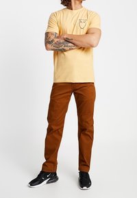 Żółta bawełniana koszulka z grafiką sowy na piersi, w zestawieniu z brązowymi bawełnianymi spodniami. Model nosi czarne buty sportowe z białymi akcentami.