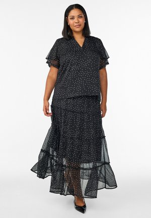 Nainen seisoo päällään musta läpikuultava kerroksellinen hame ja siihen sopiva pilkullinen pusero, jossa on röyhelöhihat, sekä mustat terävät kengät.