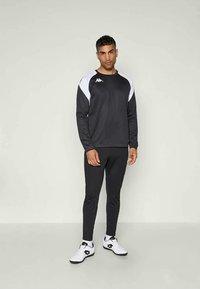 Tuta sportiva nera con accenti bianchi sulle spalle; comprende una felpa con collo tondo e pantaloni aderenti. Indossata con scarpe da ginnastica bianche.
