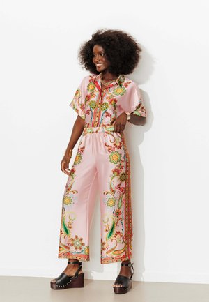 Femme souriante avec afro portant un chemisier rose à motifs floraux et un pantalon large, sandales plateformes noires, debout contre un mur blanc.