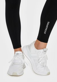 White athletic shoes with textured fabric, lace-up closure, and three-stripe design. Black fitted leggings with "SQUATPROOF" text near hem.