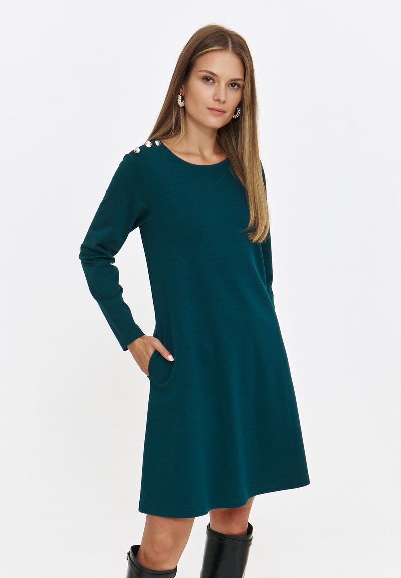 Turkusowa dzianinowa sukienka z długimi rękawami, okrągłym dekoltem, kieszeniami po bokach oraz guzikami na ramionach. Gładka faktura, sylwetka w kształcie litery A.