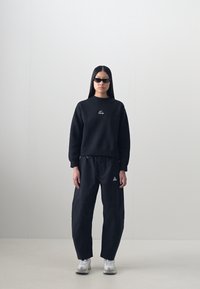 Black sweatshirt with a round neck and logo, paired with black loose-fitting trousers featuring a triangular emblem and white sneakers.