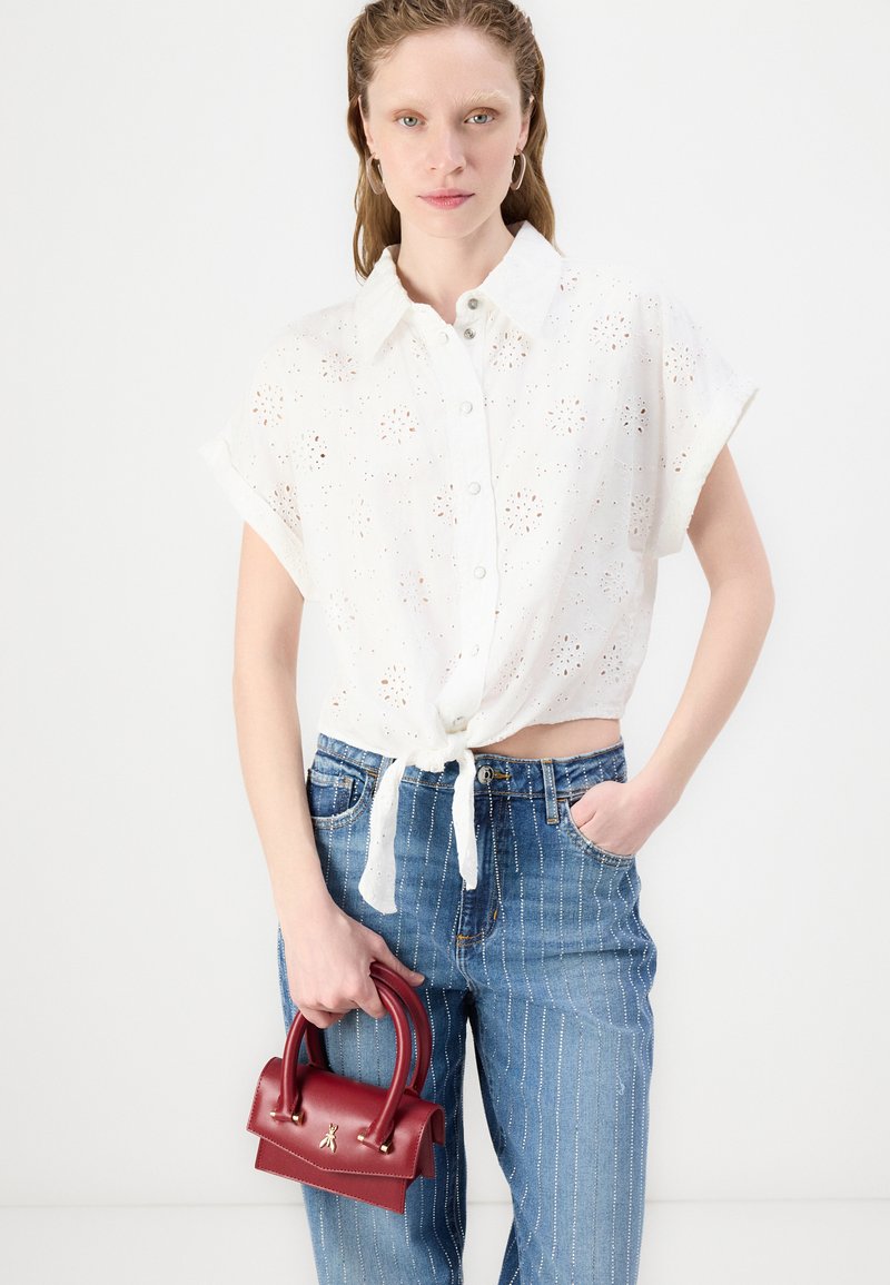 Camisa blanca de algodón con patrón de ojales, mangas cortas enrolladas, anudada en la cintura. Combinada con jeans de rayas azules y sosteniendo un pequeño bolso rojo.