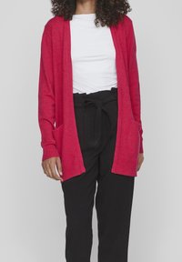 Cárdigan de manga larga y apertura frontal en un vibrante tono rojo, confeccionado en una mezcla de lana, que cuenta con dos bolsillos frontales y puños acanalados. Combinado con una camiseta blanca y pantalones negros.