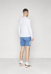 Witte sportieve long-sleeve shirt gecombineerd met blauwe shorts met drie verticale gele strepen; model draagt lichtgekleurde voetbalschoenen.