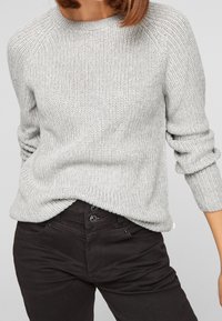 Grauer Strickpullover mit strukturierter Oberfläche und Raglanärmeln, kombiniert mit eng anliegenden schwarzen Hosen mit Knopfverschluss.