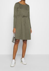 Oliwkowa sukienka z długim rękawem, okrągłym dekoltem iściągaczem w talii. W połączeniu z białymi trampkami i małą metaliczną torebką.