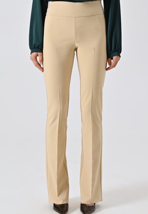 Donna che indossa pantaloni beige su misura e una blusa verde scuro a maniche lunghe, in piedi con le mani rilassate ai lati, scarpe nere a punta visibili.