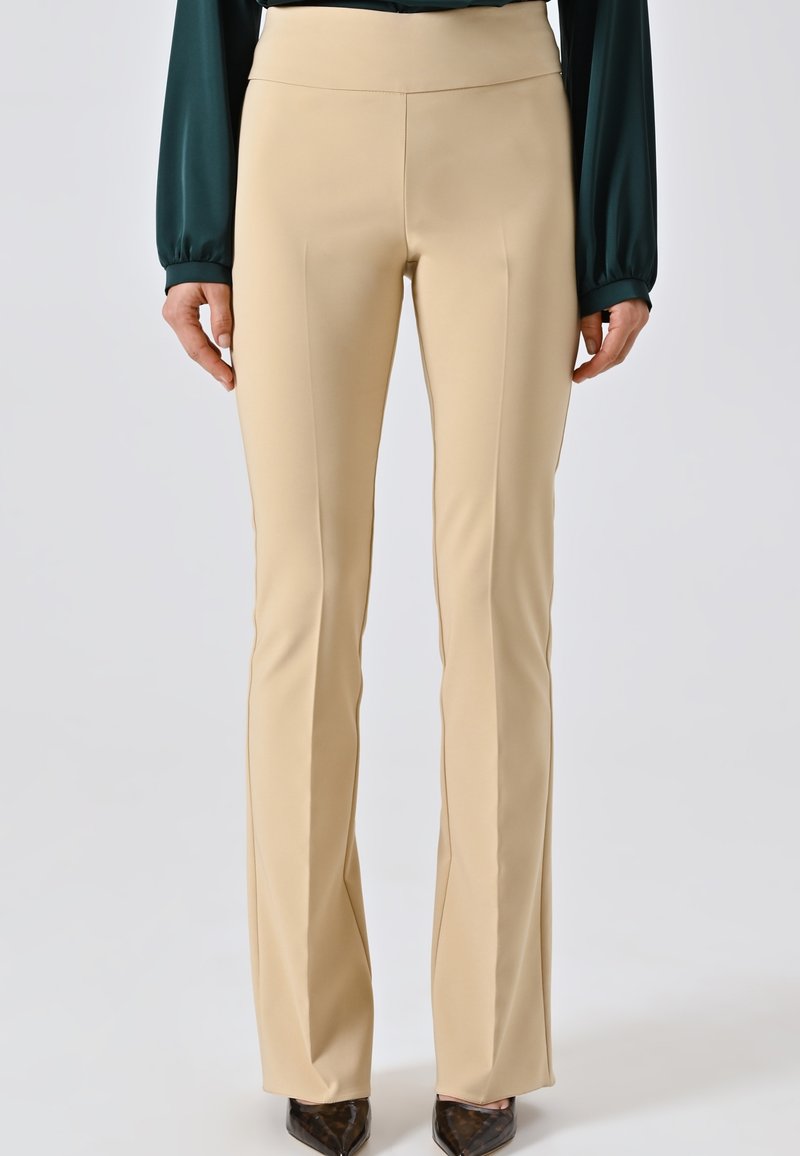Donna che indossa pantaloni beige su misura e una blusa verde scuro a maniche lunghe, in piedi con le mani rilassate ai lati, scarpe nere a punta visibili.
