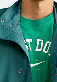 Green, lightweight jacket featuring snap fasteners, over a green t-shirt with "JUST DO IT" in white and a swoosh logo in yellow.