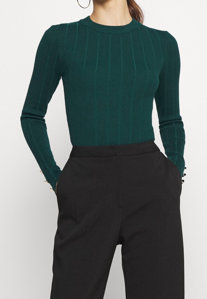 Femme portant un pull côtelé moulant vert foncé à manches longues et un pantalon noir taille haute, mains dans les poches, cadrée au niveau du cou et des cuisses.