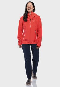 Red waterproof jacket with a front zipper, two side pockets, adjustable hood, and a slim fit. Paired with navy pants and gray shoes.