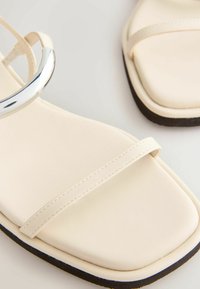 Közeli kép krémszínű szandálokról, négyzet alakú orr-résszel, vékony pántokkal és fekete szegélyű talppal világos háttéren.