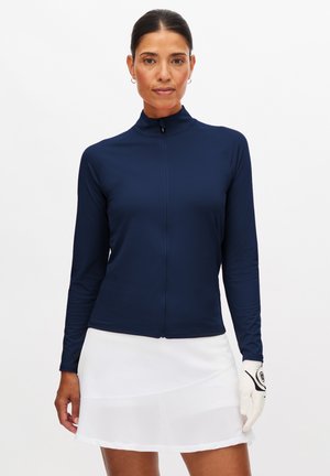 Donna che indossa una maglia blu navy con zip e maniche lunghe, gonna bianca e guanto da golf bianco sulla mano destra, in piedi davanti a uno sfondo semplice.