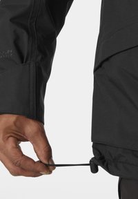 Chaqueta negra impermeable con textura lisa, que presenta un cordón en el dobladillo siendo ajustado por una mano, destacando su funcionalidad.