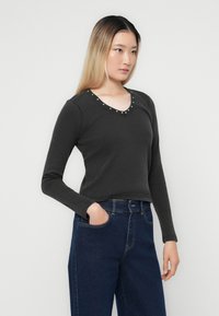 Mørk grå langermet topp med V-hals og dekorative metallnagler langs halslinjen. Ribbet tekstur, kombinert med høytlivde blå denimjeans.