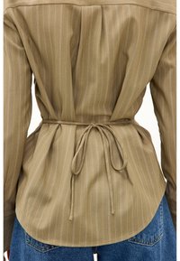 Vue de dos d'une chemise beige à fines rayures avec une ceinture fine à nouer à la taille, associée à un jean en denim bleu.