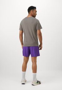 Graues Baumwoll-T-Shirt und lila Sportshorts, stehende Figur von hinten betrachtet. Weiße Crew-Socken und mehrfarbige Sneaker runden das Outfit ab.