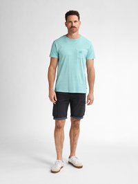 T-shirt in cotone color teal chiaro con una piccola tasca sul petto, abbinato a pantaloni cargo scuri e sneakers bianche. Design semplice e casual.