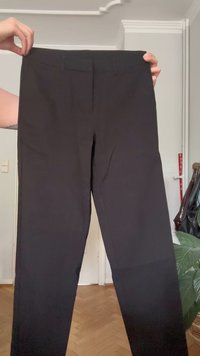 Handen houden een paar zwarte, rechte broeken omhoog in een kamer met een houten vloer en een plant in de buurt.