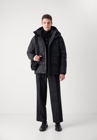 Zwarte pufferjas met een capuchon, voorzien van twee frontzakken en een ritssluiting. Draag het met een zwart shirt en op maat gemaakte broek. Zwarte schoenen.