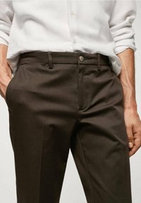 Osoba v hnědých kalhotách a bílé košili, stojící s rukama položenýma v kapsách, předvádějící upravený outfit.