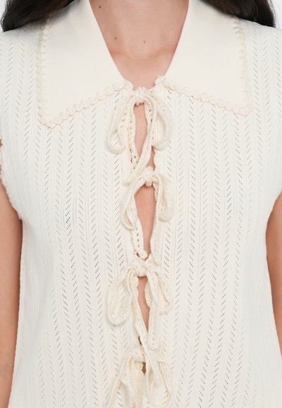 Haut sans manches en tricot crème avec un col pointu, présentant une ouverture en forme de clé ornée de nœuds texturés et d'un motif en chevrons.