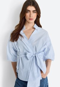 Helleblau gestreifte Bluse mit kurzen Ärmeln, einem Wickeldesign und einem vorderen Schleifenakzent. Hergestellt aus weichem Stoff mit glatter Textur.