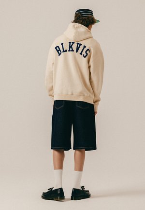 Person trägt beigen Kapuzenpullover mit dem Schriftzug "BLKVIS" auf dem Rücken, dunkle knielange Shorts, weiße Socken, schwarze Schuhe und eine schwarze Kappe mit weißen Streifen.