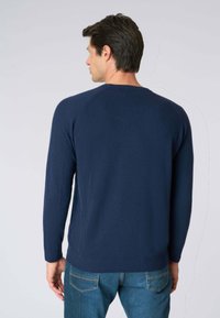 Maglione blu navy lavorato a maglia con collo rotondo, dotato di polsini e orlo a costine e dettagli di cucitura discreti lungo i lati. Indossato con jeans blu.