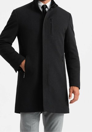 Hombre con abrigo negro hasta la rodilla sobre camisa blanca, corbata negra y pantalones grises, con una mano en el bolsillo del abrigo.