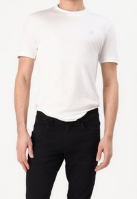Camisa de algodón blanca de manga corta, con cuello redondo, que presenta un pequeño logo azul claro en el pecho. Combinada con vaqueros negros.