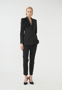 Svart skräddarsydd blazer med blommönsterbroderi på axeln, i kombination med slim-fit svarta byxor och svarta högklackade skor.