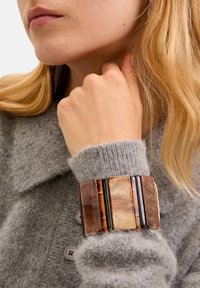 Bracciale multi-strato marrone e ambra con segmenti rettangolari, indossato su un maglione grigio in stile peloso, con scollatura a collare e dettaglio dei bottoni.
