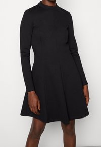 Kvinna som bär en långärmad, svart klänning knälång med en åtsittande överdel och utsvängd kjol mot en enfärgad bakgrund.