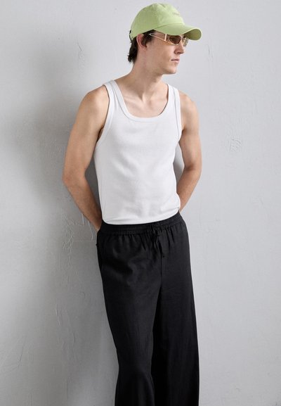 Top sin mangas de algodón blanco combinado con pantalones negros con cordón. El modelo lleva una gorra verde claro y gafas de sol doradas, de pie frente a un fondo gris.