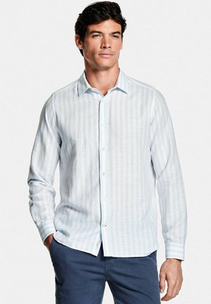 Homme portant une chemise à rayures verticales bleu clair et un pantalon bleu foncé, debout avec une main dans la poche devant un fond blanc.