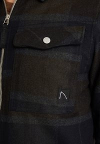 Kapsa na tmavém károvaném sáčku s klopou zabezpečenou stříbrným cvokem a malou černou nášivkou s bílým minimalistickým logem.