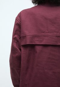 Burgunderjakke laget av tykk stoff med en strukturert design. Har en bakstykke og synlige sømmer på ermene og lommene.